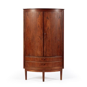 May include: A tall, curved corner cabinet made of dark brown wood with a prominent grain pattern. The piece features two vertical doors on the upper section and two horizontal drawers below, all supported by four slender, tapered wooden legs.