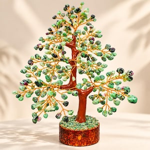 May include: A decorative tree sculpture featuring a brown trunk and branches made of thin gold-colored wire. The branches are adorned with numerous small, tumbled green and magenta gemstone chips. The base is a cylindrical brown wooden block.