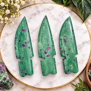 Pode incluir: Três esculturas de pedra polida em forma de pontas de flecha ou lâminas, feitas de zoisite verde com cristais de rubi magenta incrustados. Cada peça apresenta uma ponta, uma superfície lisa e uma base entalhada, exibindo variações minerais naturais.
