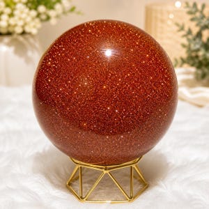 May include: A large, round, sparkling, reddish-brown sphere, possibly a decorative crystal ball, sits on a gold-colored geometric metal stand. The sphere has a polished surface with many tiny, reflective inclusions, giving it a glittery appearance.