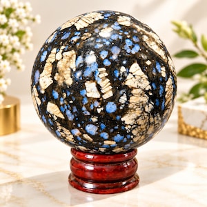 Puede incluir: Una piedra esférica pulida que presenta un patrón complejo de inclusiones minerales en negro profundo, azul vibrante y blanco crema. La esfera está montada sobre una base circular de madera de color rojo oscuro brillante, con una superficie lisa y reflectante.