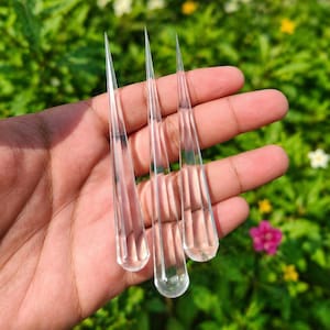 Könnte beinhalten: Drei klare, tropfenförmige Glasobjekte mit spitzen Spitzen werden in einer Hand gehalten. Die Objekte sind transparent, reflektieren Licht und scheinen dekorative oder funktionale Gegenstände zu sein. Der Hintergrund ist eine verschwommene Ansicht von grünem Laub.