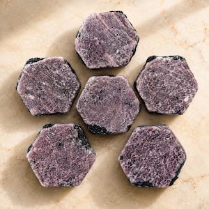 May include: Six hexagonal raw ruby crystals arranged in a circular pattern. Each stone features a deep purple-red hue with a textured, crystalline surface and dark, rough edges. The stones display natural mineral formations with visible internal fractures.