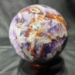 May include: A polished spherical stone featuring a mix of deep purple amethyst and translucent white quartz sections, intersected by prominent veins of reddish-brown iron oxide inclusions throughout the entire surface of the orb.