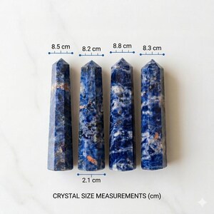 Op de afbeelding: Vier blauwe sodaliet kristallen obelisken, elk met een puntige top. Afmetingen: 8,5 cm, 8,2 cm, 8,8 cm en 8,3 cm hoog, met een breedte van 2,1 cm. De kristallen hebben een gemarmerd uiterlijk met witte en oranje insluitsels.