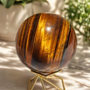 Pode incluir: Uma esfera polida de olho de tigre, exibindo faixas de marrom dourado e preto. A pedra preciosa é exibida em um suporte geométrico dourado, criando um contraste marcante. A superfície da esfera reflete a luz.