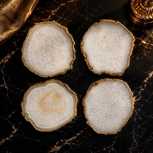May include: A set of four circular coasters made of translucent white agate stone. Each coaster features a natural, irregular crystalline texture and is finished with a metallic gold-colored electroplated edge that follows the organic shape.