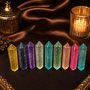 May include: A collection of nine colorful crystal wands in shades of yellow, blue, purple, green, and pink. The crystals are arranged on a dark surface with a gold-colored decorative container and a candle holder in the background.