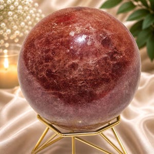 Può includere: Una grande sfera di cristallo lucidata, di colore rosso-viola. La sfera è esposta su un supporto in metallo dorato con una base a tre gambe. Il cristallo ha una superficie strutturata e varie sfumature di rosso e viola.