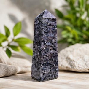 May include: A polished stone obelisk featuring a multifaceted, crystalline texture with deep shades of dark blue, charcoal grey, and black. The tower has a pointed, pyramidal top and a rectangular base, showcasing natural mineral inclusions throughout.