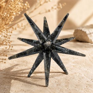 Pode incluir: Uma escultura em forma de estrela com múltiplas pontas, esculpida em pedra labradorite cinzento-escura. O objeto apresenta doze pontas afiadas que irradiam de um núcleo central, exibindo a textura cristalina natural e as inclusões minerais.