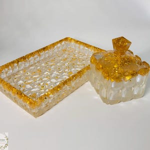 May include: A decorative tray and a small lidded box. Both are clear with a textured surface and gold glitter accents. The tray is rectangular, and the box is square with a geometric pattern.
