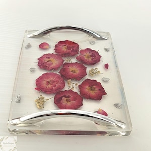 May include: A rectangular clear resin tray with embedded dried red roses and silver-colored accents. The tray has two curved, silver-colored handles. The roses are arranged in a cluster, with small dried yellow flowers and silver-colored stones scattered around them.