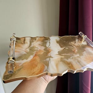 May include: A rectangular decorative tray with a wavy edge. The tray features a gold, white, and orange marbled design. It has clear acrylic handles with gold accents. The tray is held by a person.