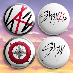 May include: Four white button pins with different designs. One pin has the word "LAZ" in black with a red cross. Another pin has the words "Stray Kids" in black and red. A third pin has a red and silver compass design with the words "Stray Kids". The last pin has the word "Stay" in black.