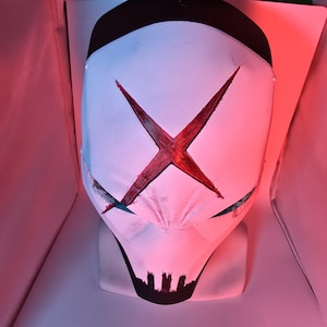 May include: A white full-face mask featuring a prominent red X symbol across the forehead and eyes. The mask has a black top section and a black jagged design at the chin area. The surface shows a textured finish with dark accents around the eyes.