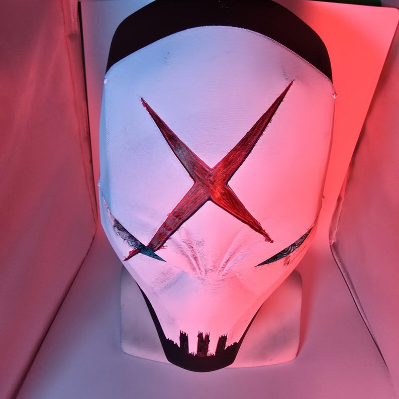 May include: A white full-face mask featuring a prominent red X symbol across the forehead and eyes. The mask has a black top section and a black jagged design at the chin area. The surface shows a textured finish with dark accents around the eyes.
