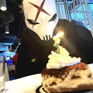 May include: A person wearing a white mask with a red X symbol and black eye markings, looking at a slice of tiramisu topped with whipped cream and a single lit candle. The mask features a skull-like design with dark vertical lines at the chin.