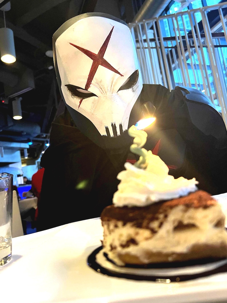 May include: A person wearing a white mask with a red X symbol and black eye markings, looking at a slice of tiramisu topped with whipped cream and a single lit candle. The mask features a skull-like design with dark vertical lines at the chin.