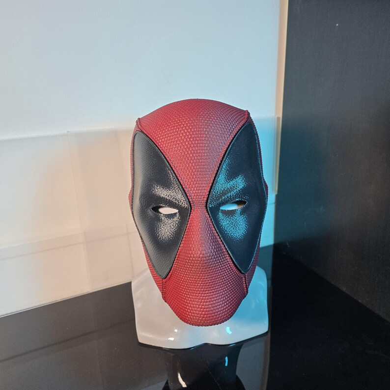 Puede incluir: Una m&aacute;scara de Deadpool roja y negra con una superficie texturizada. La m&aacute;scara tiene una m&aacute;scara para los ojos negra con ojos blancos.