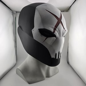 May include: A full-face helmet featuring a matte black back and a white front panel. The white section displays a prominent dark red X symbol on the forehead, narrow black eye openings, and a series of vertical black slits at the mouth area.