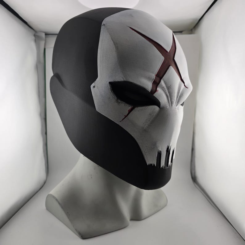 May include: A full-face helmet featuring a matte black back and a white front panel. The white section displays a prominent dark red X symbol on the forehead, narrow black eye openings, and a series of vertical black slits at the mouth area.