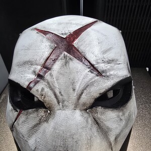 May include: A white full-face mask featuring a weathered, distressed finish with black shading. A prominent, deep red X-shaped symbol is centered on the forehead. The mask includes two large, angular black eye openings and sculpted facial contours.