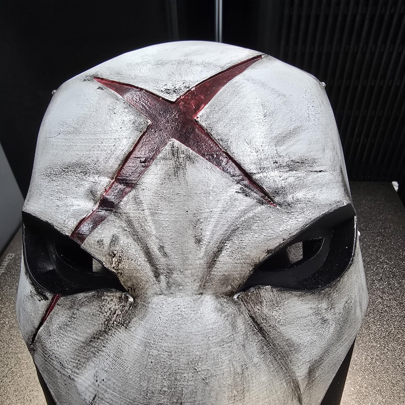 May include: A white full-face mask featuring a weathered, distressed finish with black shading. A prominent, deep red X-shaped symbol is centered on the forehead. The mask includes two large, angular black eye openings and sculpted facial contours.