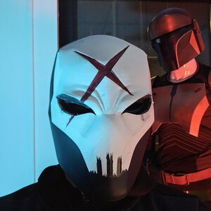 May include: A white mask with a dark red X symbol on the forehead and black markings around the eyes and mouth. In the background, a dark helmet with a T-shaped visor and orange accents is visible on a mannequin torso.