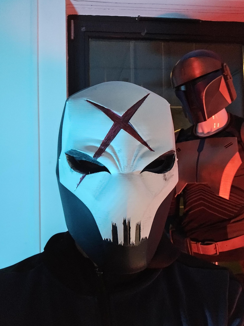 May include: A white mask with a dark red X symbol on the forehead and black markings around the eyes and mouth. In the background, a dark helmet with a T-shaped visor and orange accents is visible on a mannequin torso.