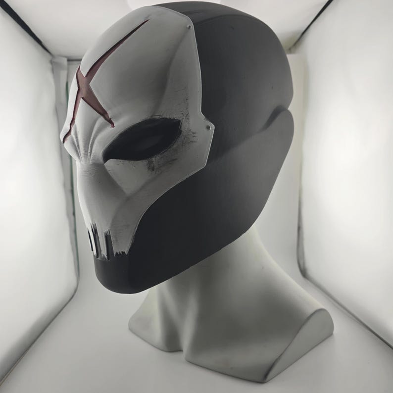 May include: A full-head helmet featuring a matte black base and a white faceplate. The faceplate displays a prominent red X symbol on the forehead, dark eye openings, and vertical ventilation slits at the chin with subtle weathered detailing.