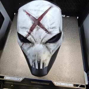 May include: A white full-face mask with a weathered, distressed finish and a prominent dark red X symbol on the forehead. The mask features deep black eye sockets, a jagged mouth area, and subtle black shading throughout the surface texture.