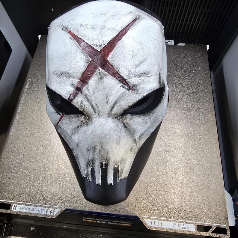 May include: A white full-face mask with a weathered, distressed finish and a prominent dark red X symbol on the forehead. The mask features deep black eye sockets, a jagged mouth area, and subtle black shading throughout the surface texture.