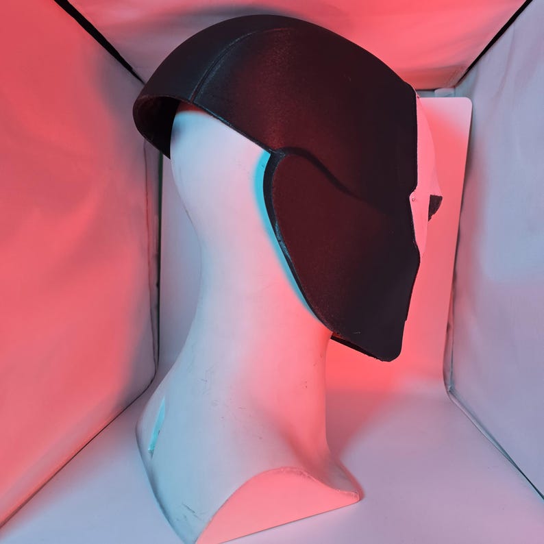 May include: A two-toned mask featuring a matte black upper section and a white lower section, displayed on a white mannequin head. The mask covers the upper face and sides, with a smooth, curved design that contours to the shape of the head.