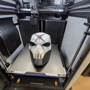 May include: A 3D printed full-face mask featuring a white upper section with a weathered finish, a prominent red X symbol on the forehead, and a black lower section with vertical grill-like slits over the mouth area, resting on a printer bed.