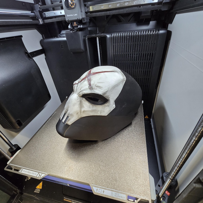 May include: A full-face helmet featuring a matte black base and a weathered white faceplate. The faceplate displays a prominent red X symbol on the forehead and vertical slits over the nose area, with dark eye openings and a distressed finish.
