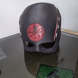 May include: A black plastic mask with a red Hydra logo. The mask features a skull and tentacles in the logo. The mask has a black visor and red accents.