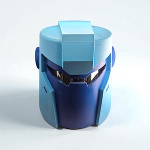 Puede incluir: Un casco azul y azul marino con un diseño futurista. El casco tiene una visera y orejeras. La parte superior del casco tiene forma rectangular. El casco está hecho de un material duro.