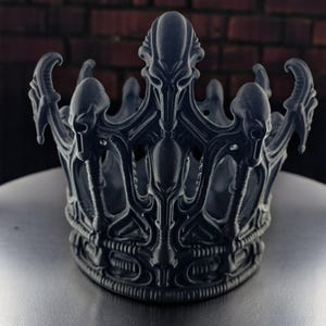 May include: A dark gray crown with an intricate, alien-inspired design. The crown features a series of stylized, skeletal-like forms and curved elements. The crown is made of a solid material and sits on a metallic surface.