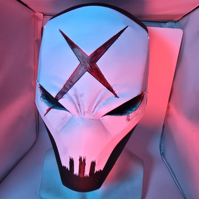 May include: A white full-face mask featuring a prominent red X symbol on the forehead, narrow angular eye openings, and a black jagged mouth design at the chin. The mask is illuminated by blue and red light, highlighting its textured surface.