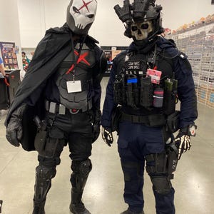 May include: Two individuals in tactical cosplay costumes. The left figure wears a white skull mask with a red X, black armor, and a cape. The right figure wears a skull mask, night vision goggles, a dark blue uniform with SAS patches, and tactical gear.