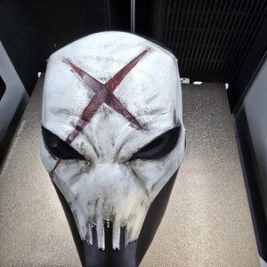 May include: A white full-face mask featuring a distressed finish with black weathering and a prominent red X symbol centered on the forehead. The mask has deep-set black eye sockets and a stylized mouth area with vertical slits.