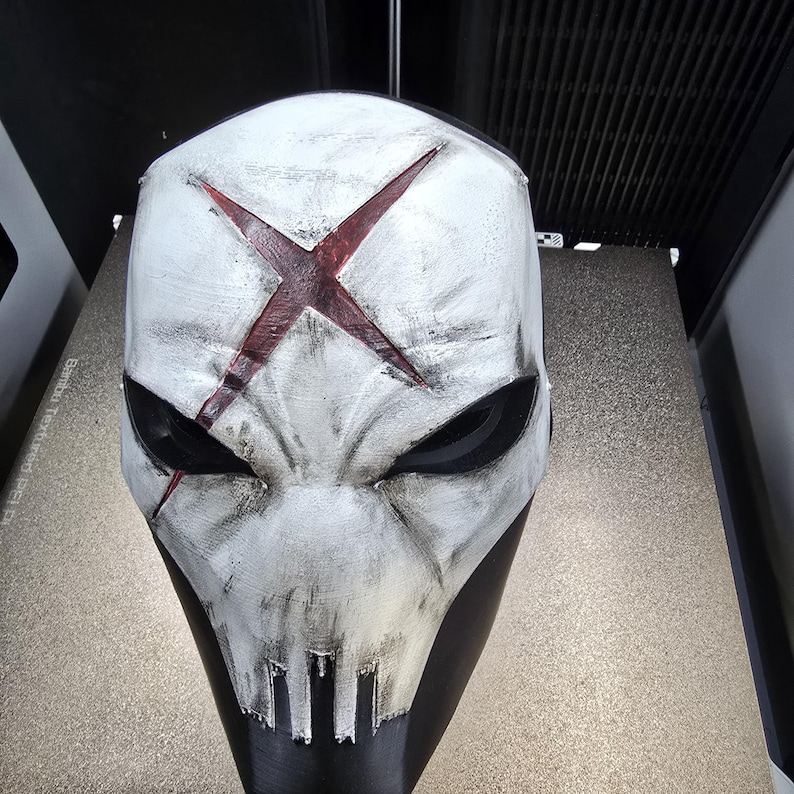 May include: A white full-face mask featuring a distressed finish with black weathering and a prominent red X symbol centered on the forehead. The mask has deep-set black eye sockets and a stylized mouth area with vertical slits.
