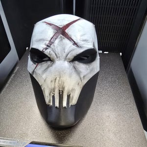 May include: A full-face mask featuring a weathered white finish with a prominent dark red X symbol on the forehead. The mask has deep black eye sockets and a jagged, skull-like mouth design, with a solid black lower section and textured surface.