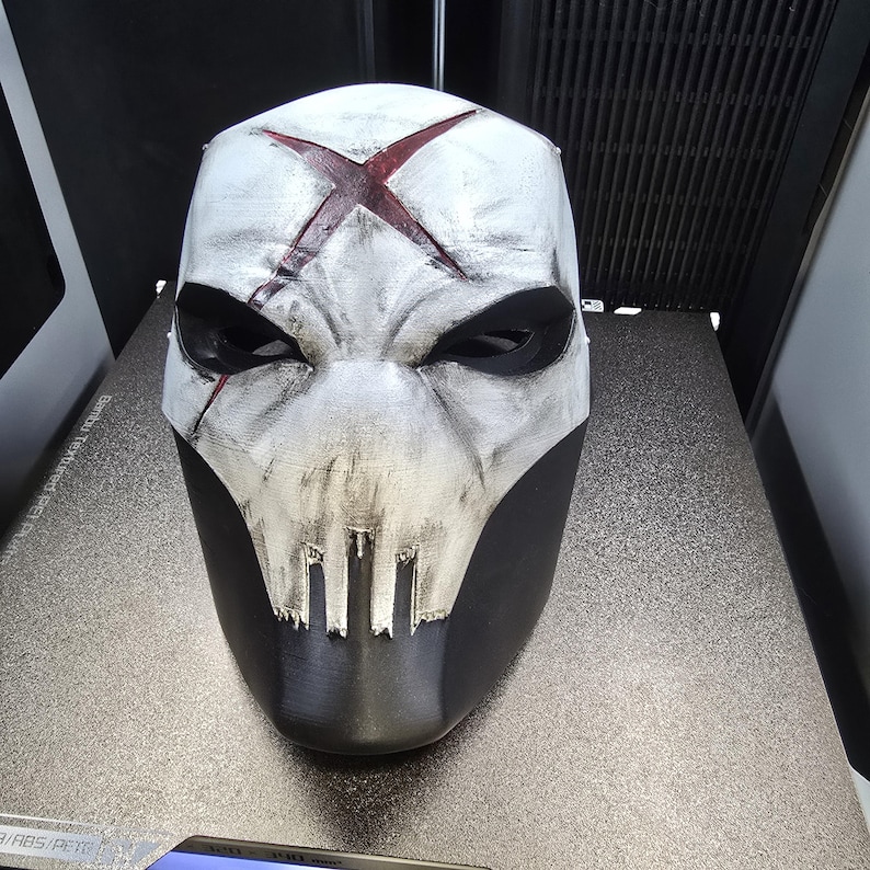 May include: A full-face mask featuring a weathered white finish with a prominent dark red X symbol on the forehead. The mask has deep black eye sockets and a jagged, skull-like mouth design, with a solid black lower section and textured surface.