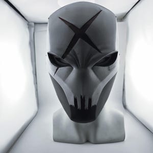 May include: A matte grey full-face mask featuring a prominent dark X-shaped cutout on the forehead, angular eye openings, and a jagged, tooth-like design along the lower mouth area, displayed on a neutral mannequin head bust.