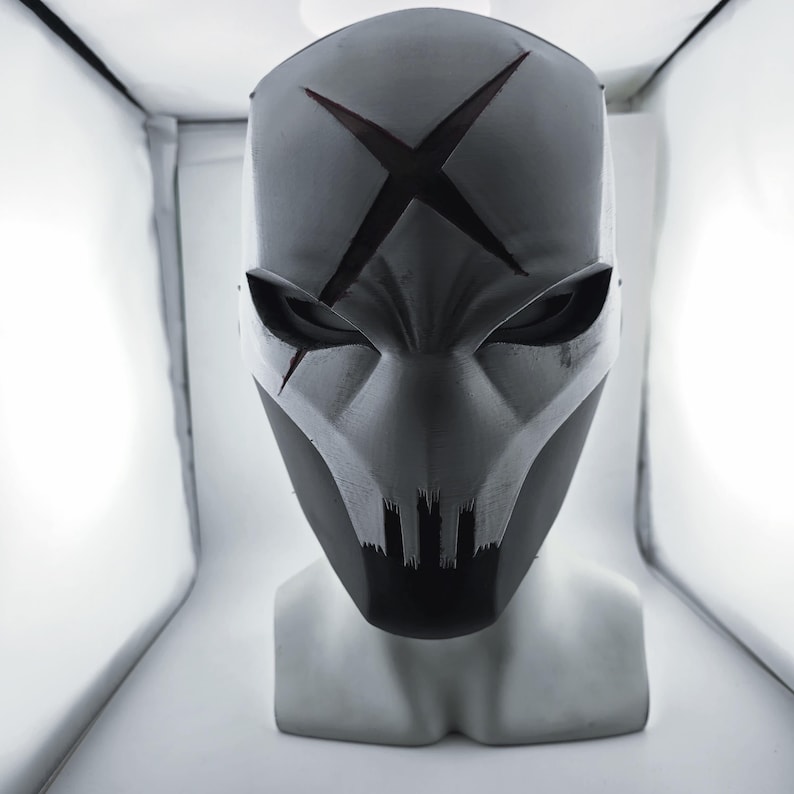 May include: A matte grey full-face mask featuring a prominent dark X-shaped cutout on the forehead, angular eye openings, and a jagged, tooth-like design along the lower mouth area, displayed on a neutral mannequin head bust.
