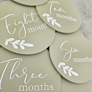 May include: A set of ten round, sage green baby milestone photo props with white text and leaves. The text on the props reads "One month", "Two months", "Three months", "Four months", "Five months", "Six months", "Seven months", "Eight months", "Nine months", and "Ten months".