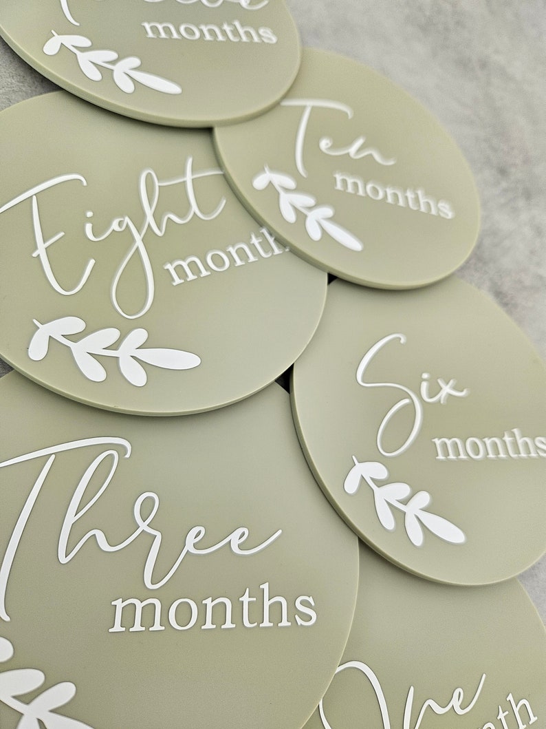 May include: A set of ten round, sage green baby milestone photo props with white text and leaves. The text on the props reads "One month", "Two months", "Three months", "Four months", "Five months", "Six months", "Seven months", "Eight months", "Nine months", and "Ten months".