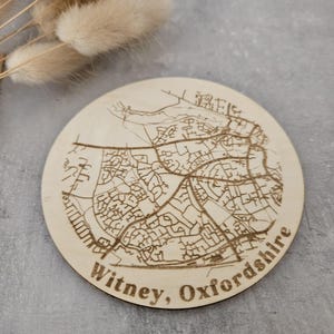May include: A round wooden coaster featuring a laser-etched map of Witney, Oxfordshire. The coaster is light brown and has a natural wood grain.